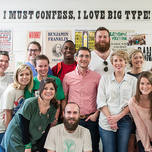 A graphic design class poses in front of typography samples, a banner above their heads reads 'I must confess, I love big type.'