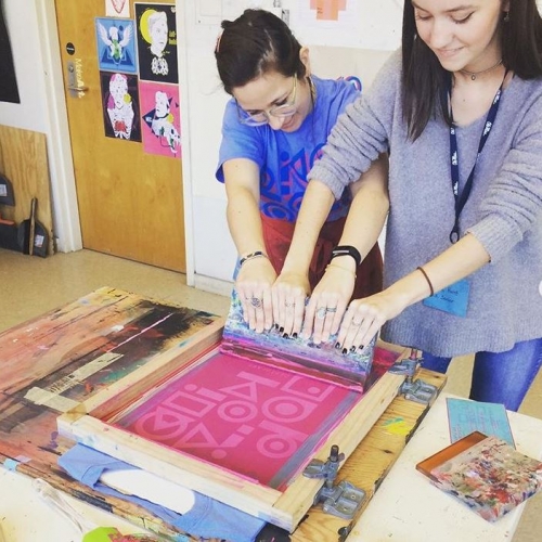 Two women working with the silk screen to create a pink design on a t-shirt