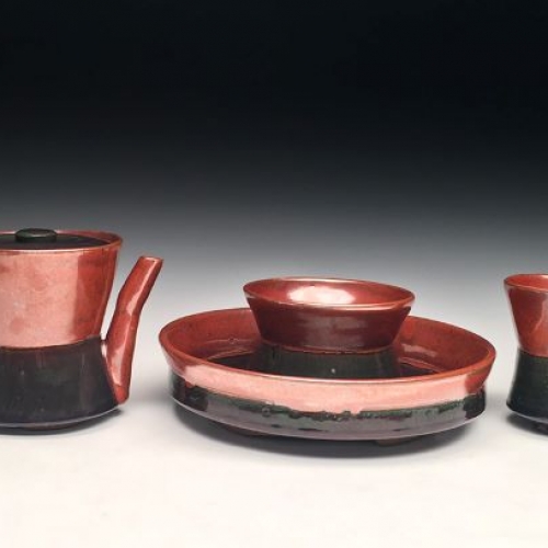 A red, shiny ceramic tea kettle, cups, and a dish