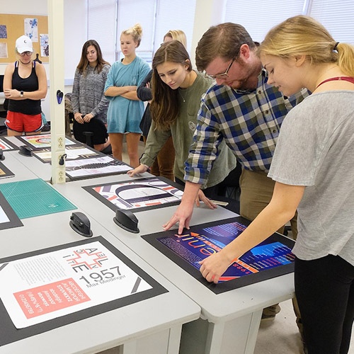 Graphic design students gathered around a table study prints of graphic art